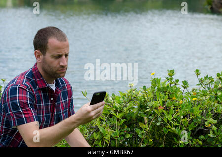 Man sitting and on a smartphone in front of a lake outdoors wearing a plaid shirt. Stock Photo