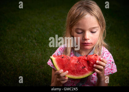 Cute little blond girl eating watermelon on the grass in summertime Stock Photo