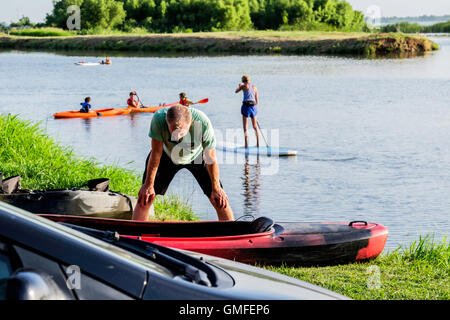 A middle aged Caucasian man looks over his kayak on shore while people play on a paddleboard and kayaks on a river. Stock Photo