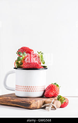 Fresh ripe red strawberries in country style enamel mug on rustic wooden board over white background