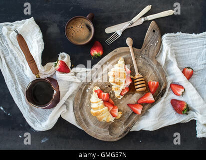 Breakfast set. Freshly baked croissants with strawberries, mascarpone, honey and coffee on rustic wooden board over dark grunge backdrop Stock Photo