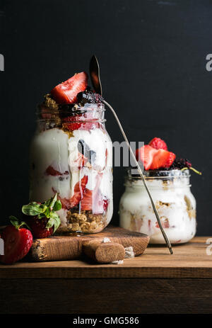 Yogurt oat granola with strawberries, mulberries, honey and mint leaves in tall glass jar on black backdrop Stock Photo