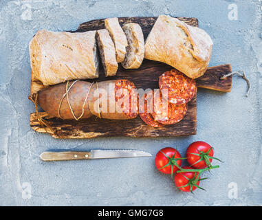 Wine snack set. Hungarian mangalica pork salami sausage, rustic bread and fresh tomatoes on dark wooden board over a rough grey-blue concrete background Stock Photo