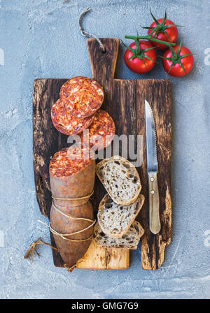 Wine snack set. Hungarian mangalica pork salami sausage, rustic bread and fresh tomatoes on dark wooden board over a rough grey-blue concrete background Stock Photo