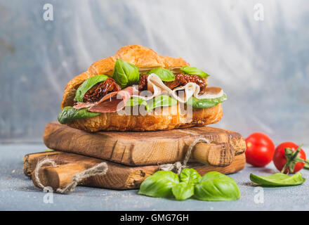 Croissant sandwich with smoked meat Prosciutto di Parma, sun dried tomatoes, fresh spinach and basil on stone textured grey background Stock Photo