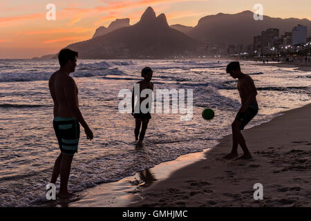 Young men play football on Ipanema beach silhouetted by sunset with the Two Brother mountains in the distance in Rio de Janeiro, Brazil.