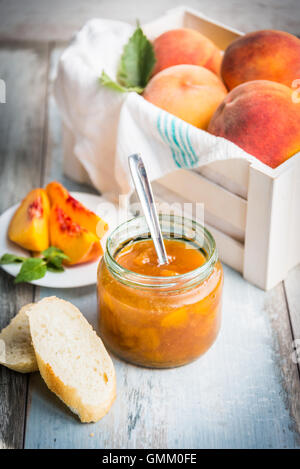A jar with homemade peach jam in front of a white wood crate full of ripe peaches over a light blue old wood background. Stock Photo