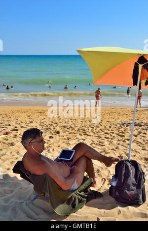 Man with tablet computer and headphones at seaside on sandy beach under umbrella in shade Stock Photo