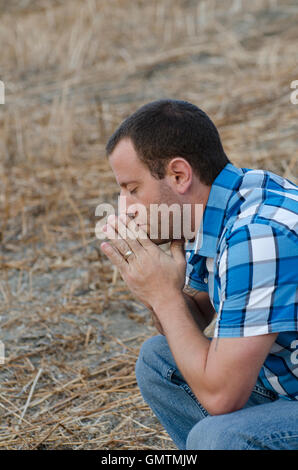 Man crouching down and praying with his hands together on a hillside wearing a plaid shirt. Stock Photo