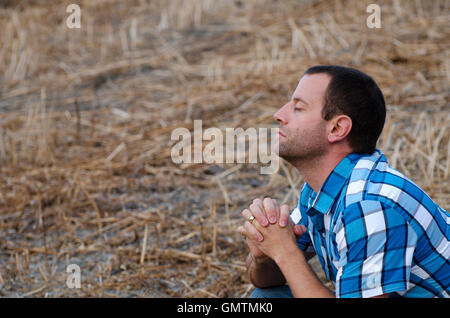 Man crouching down and praying with his hands clasped on a hillside wearing a plaid shirt. Stock Photo