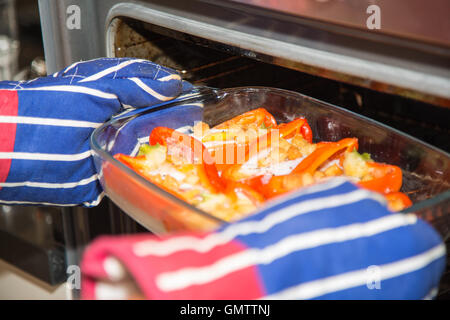 Cooked stuffed red peppers with eggs, ham and croutons in an oven proof serving dish being taken from an oven with red and blue Stock Photo
