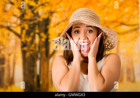 Young attractive woman wearing white top and fashionable hat, touching cheeks with own hands screaming out, forest autumn background Stock Photo