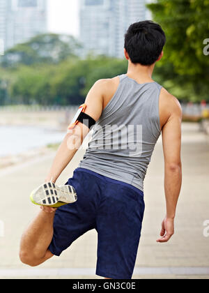 young asian male jogger with fitness tracker attached to arm warming up by stretching leg before running. Stock Photo