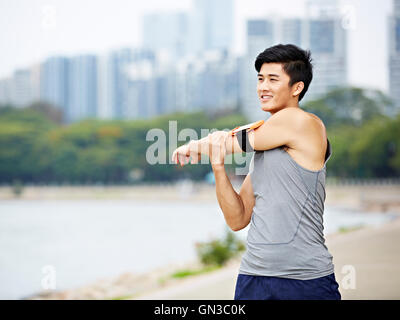 young asian male jogger with fitness tracker attached to arm warming up by stretching arms and upper body before running, city s Stock Photo