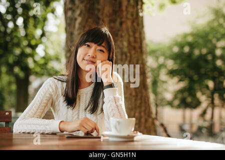 Shot of happy young woman sitting at cafe table with digital tablet and cup of coffee. Chinese female model relaxing at outdoor Stock Photo