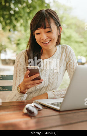 Shot of smiling asian woman using cellphone at outdoor coffee shop. Female sitting at cafe table outdoors using smartphone. Stock Photo