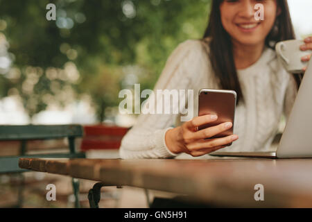 Young woman in a cafe reading a text message from her mobile phone. Chinese female sitting at cafe table with laptop and using s Stock Photo