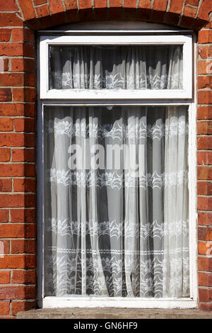 old net curtains hanging in the window of a red brick victorian terraced house Stock Photo