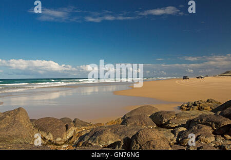 Vast 75-mile beach, ocean & rocks at Indian Head on Fraser Island with distant vehicles on highway, blue sky reflected in water Stock Photo