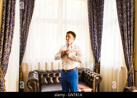 Handsome man putting on shirt standing near window at his room in morning. New opportunities, dating, wedding day or getting ready for job interview concept Stock Photo