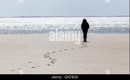 A silhouette of a young woman stands on a beach, leaving her foot prints in the sand and a lighthouse in the distance. Stock Photo