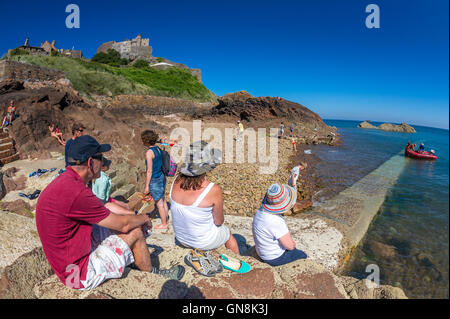 Holiday makers sit on concrete steps at Gorey harbour with Mont Orgueil castle in the background, Jersey. Stock Photo