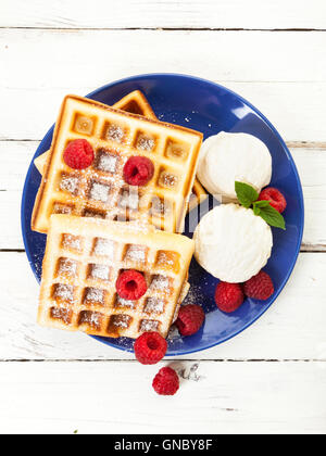Belgian waffles with raspberries, vanilla ice cream scoops and powdered sugar on blue plate, on rustic wooden table, top view Stock Photo