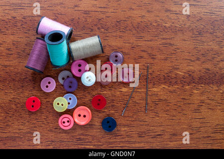 spools of cotton thread, a selection of different colored buttons and sewing needles on a wooden darning table top Stock Photo
