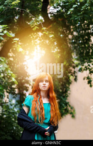 Young trendy boho woman with red head posing in green park. Sunlight effect. Stock Photo