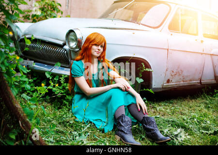 Funny bright young woman posing near vintage abandoned old car. Full body portrait. Smiling and sitting in grass. Colored red lo Stock Photo