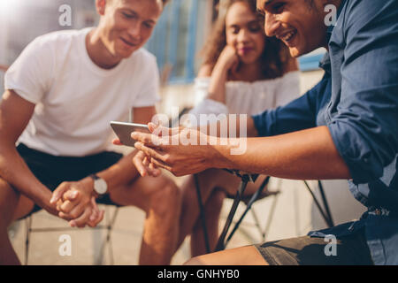 Young people sitting outdoors and looking at mobile phone. Group of friends sitting at outdoor cafe and watching video on the sm Stock Photo