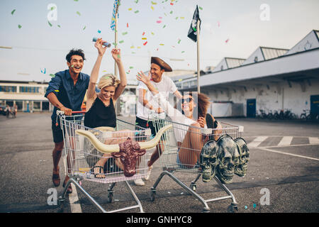 Young friends having fun on shopping trolleys. Multiethnic young people racing on shopping cart. Stock Photo