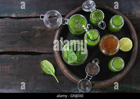 Opened glass jars and bottle of green spinach smoothie, served with baby spinach leaves, chia seeds, honey and lime in dark clay Stock Photo