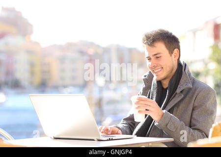Elegant handsome man using a laptop and searching information on line in a coffee shop terrace of a port with the sea Stock Photo