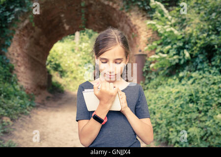 Small teenage girl with smartwatch drawing in notebook near beautiful old arch bridge in park. Cute hispanic travelling girl wri Stock Photo