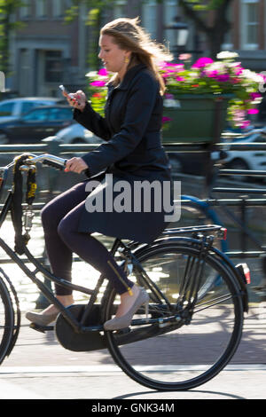 Young woman wearing high heels shoes, carrying iPhone cycling out for the evening across bridge in Amsterdam, Holland Stock Photo