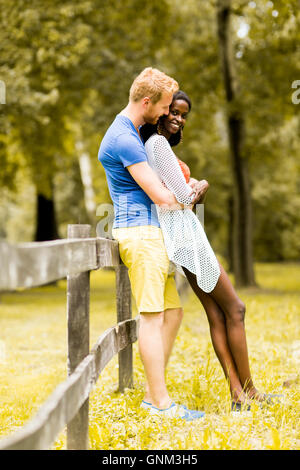 Young multiracial couple in love standing next to the fence in the park on a sunny day Stock Photo