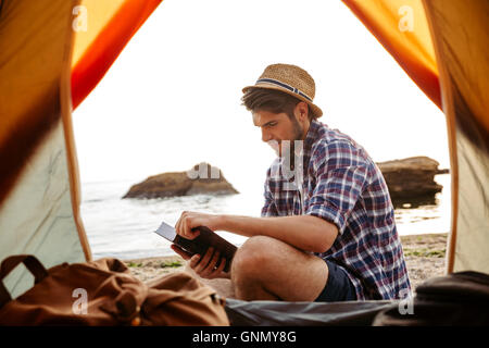 Smiling young man sitting near touristic tent and reading book at the beach Stock Photo