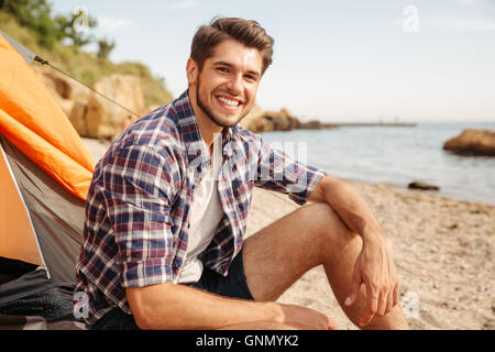 Smiling young man tourist sitting in touristic tent at the beach and looking at camera Stock Photo