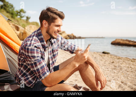 Smiling young man tourist using smartphone sitting in touristic tent at the beach Stock Photo