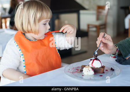 two years old kid eating with orange plastic spoon a piece of chocolate cake with vanilla ice cream sharing with woman at restau Stock Photo