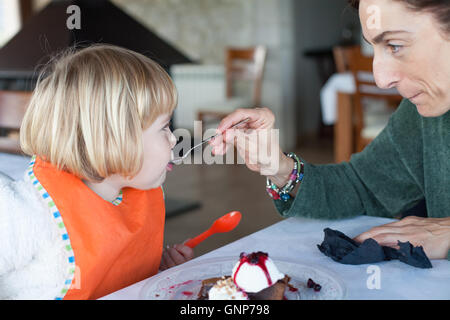 green sweater woman feeding little kid orange bib with metal spoon a piece of chocolate cake with vanilla ice cream at restauran Stock Photo