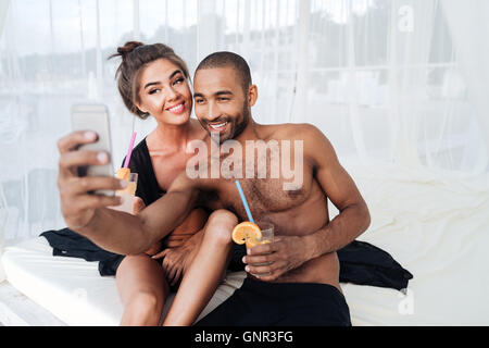 Happy smiling multiracial couple making selfie and holding cocktails at the beach Stock Photo