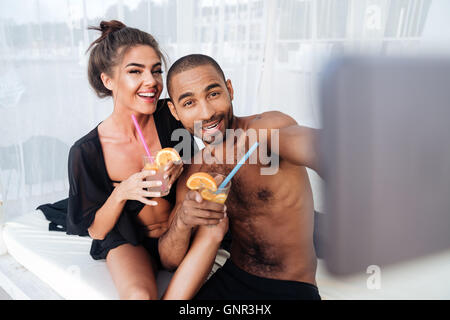 Happy smiling multiracial couple making selfie and holding cocktails at the beach Stock Photo