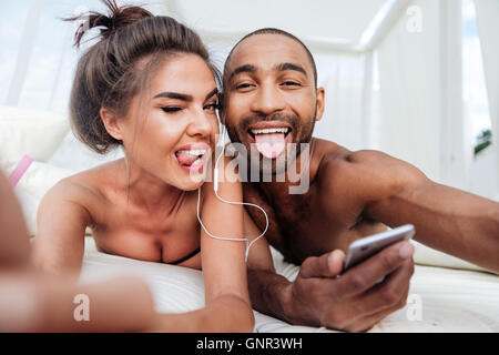 Happy cheerful couple making selfie and showing tongues while listening music at the beach Stock Photo