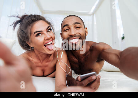 Happy cheerful couple making selfie and showing tongues while listening music at the beach Stock Photo