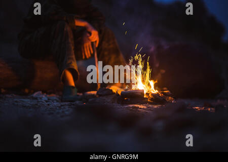 Hiker warms their feet next to a campfire at dusk camping in the woods Stock Photo