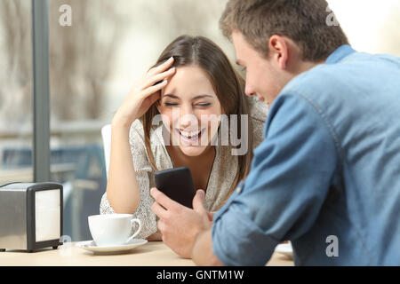 Cheerful couple laughing watching media in smart phone sitting in a coffee shop Stock Photo