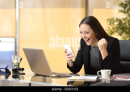Excited businesswoman winning after achievement reading a smart phone sitting in a desktop at office Stock Photo