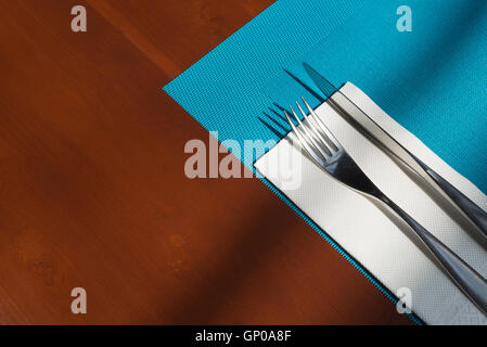 Silverware and tissue and blue mat napery on brown wooden table. Simple table setting in restaurant with light and shadow. Stock Photo